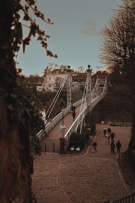 A view of the Hammersmith Bridge during daylight hours, with several pedestrians walking across the suspension bridge. The bridge features tall, white cable supports and a pedestrian walkway with a metal railing on each side. In the foreground, a cobbled pathway descends from a street level, with a few people walking and some trees without leaves on either side indicating late autumn or winter. Behind the bridge, residential buildings with classical architecture are visible on a hillside, and the sky is partly cloudy with soft natural lighting. This scene illustrates the typical setting for home relocation or furniture transport activities that Hammersmith Removals may facilitate, emphasizing the environment and logistical considerations involved in house removals near iconic London landmarks.