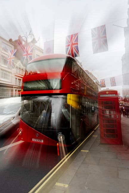 A red double-decker bus displaying 'Hammersmith' as its destination drives along a street in Hammersmith, W6, during daytime with overcast weather. The bus is parked close to the pavement, where a traditional red British telephone booth stands nearby. Above the street, decorative Union Jack flags hang across the roadway, adding a festive or patriotic touch. Behind the bus, multi-storey buildings with classic architectural features are visible, indicating a busy urban environment. The scene showcases elements typical of central London travel and urban life, relevant to home relocation and furniture transport services operated by Hammersmith Removals, as part of the area’s moving logistics. Bright reflections on the bus and street suggest daylight, while the setting appears clean and well-maintained, perfect for supporting house moving activities and logistical planning.