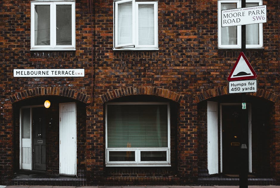 A close-up view of a brick residential building on Melbourne Terrace, SW6, featuring two arched doorways and a large ground-floor window with horizontal blinds. To the right, a street sign indicates the location as Moore Park Road SW6, and a road sign warns drivers about Humps for 450 yards. Above the doorways, two upper-floor windows with white frames and partially drawn blinds are visible. A small, round outdoor light illuminates the left doorway, which is partially open and reveals a glimpse of the interior. The building's brickwork is reddish-brown with darker mortar, and the scene is lit by natural and ambient lighting. This setting reflects the typical environment where professional house removals and furniture transport services, such as those provided by Hammersmith Removals, might prepare for the packing and loading process during a flat move or home relocation in the W6 area.