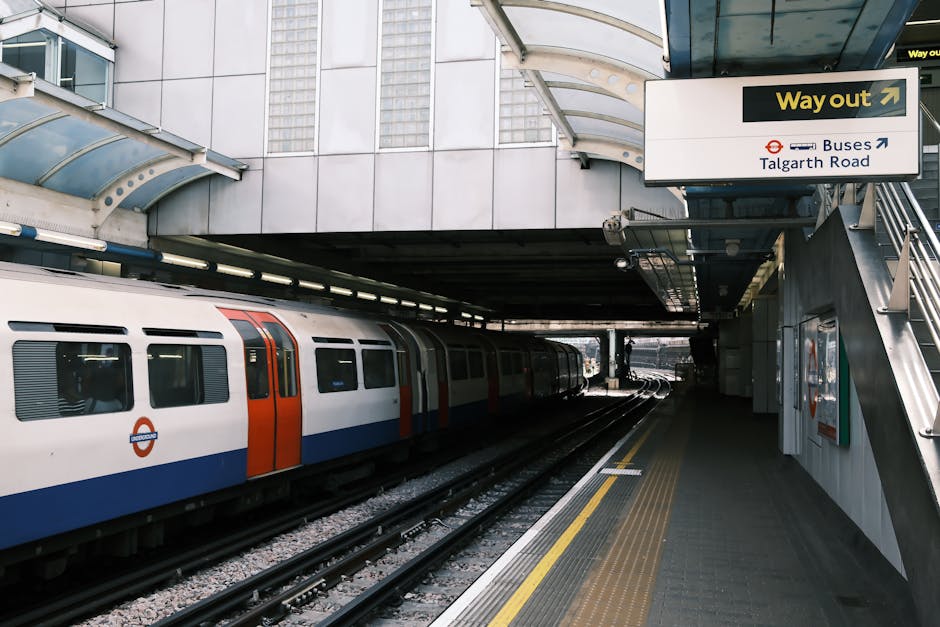 The image depicts a partially covered train station platform with a modern London Underground train stopped alongside. The train features a white body with red and blue accents, and several windows are visible along its length. The platform surface is paved with textured tiles, including a yellow tactile warning strip near the edge for safety. Overhead, a curved transparent canopy provides shelter for waiting passengers, supported by metal framing. A signboard directs passengers to the exits and bus connections, with text indicating 'Way out' and routes to Talgarth Road and bus stops. To the right, a staircase with metal railings ascends to an upper level. The station environment suggests an urban setting with a focus on efficient public transport and convenience, aligning with services like removals for home relocation by Hammersmith Removals, especially during busy moving days involving transport via train and planning for logistics.