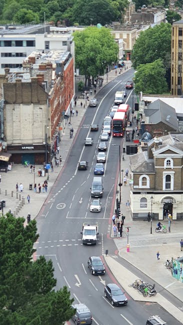 An aerial view of a busy residential street with parked cars along the curb and moving vehicles on the road, including a red double-decker bus in the distance. The sidewalk is populated with pedestrians, and several bicycles are parked near a small building at the corner. Trees with lush green foliage line the street, providing shade to nearby buildings, some of which have brick and plaster facades. The scene depicts a typical urban environment suitable for home relocation or furniture transport, with the street serving as a route for moving services such as those offered by Hammersmith Removals during a house move or packing and loading process.