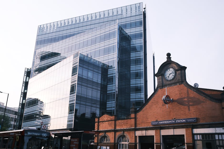 A modern urban scene in Hammersmith showing a glass-fronted office building with reflective windows towering above a historic brick building featuring a clock tower and an arched entrance. The clock on the brick structure displays the time, and below it, a sign indicates 'Hammersmith Station.' In the foreground, part of a bus is visible, along with street signs and a pavement area. This setting illustrates the typical environment for house removals and furniture transport services, with the juxtaposition of contemporary commercial architecture and traditional railway station architecture. The scene is well-lit with natural daylight, highlighting the clear glass reflections and brick textures, and captures the logistics process involved in home relocation, including the potential for loading and unloading activities near the station while [COMPANY_NAME] manages packing and moving operations efficiently.